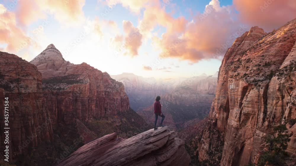 Adventurous Woman at the edge of a cliff is looking at a beautiful landscape view in the Canyon during a vibrant sunset. Taken in Zion National Park, Utah, United States. Parallax Panorama