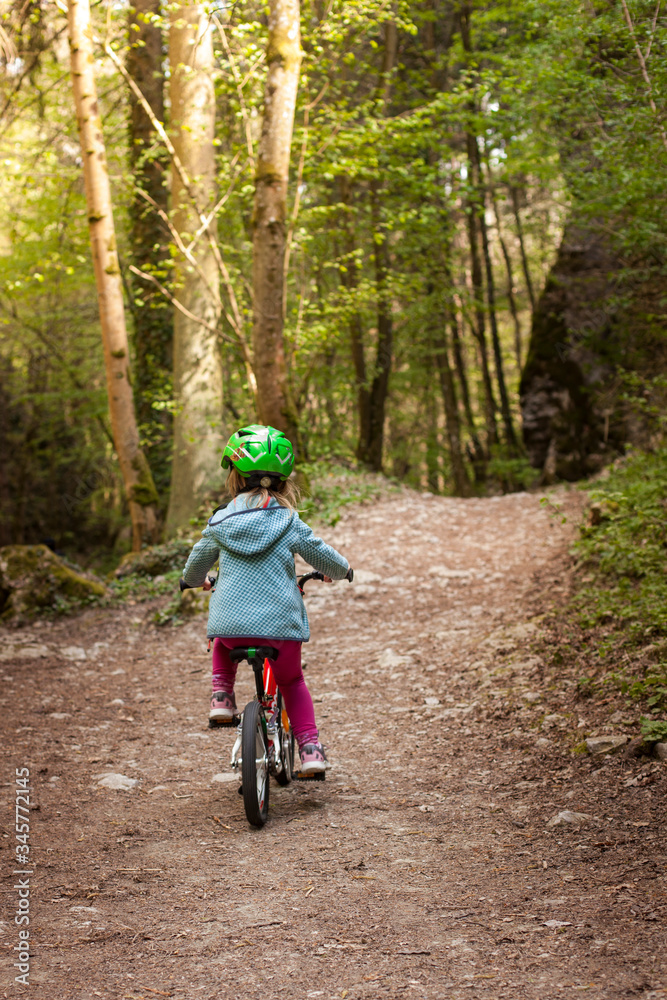 Naklejka premium Young girl with green helmet cycling on forest path. Junges Mädchen mit grünem Helm fährt mit Fahrrad auf Waldweg.