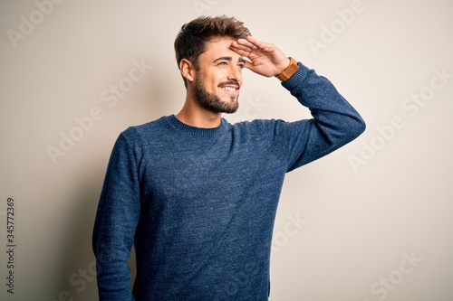 Young handsome man with beard wearing casual sweater standing over white background very happy and smiling looking far away with hand over head. Searching concept.