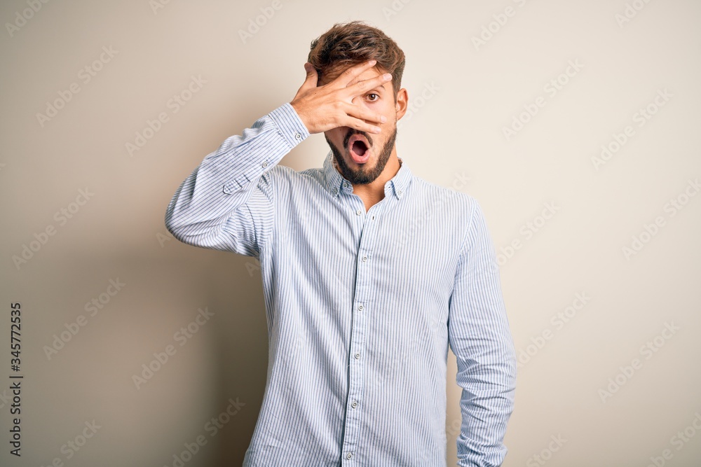 Young handsome man with beard wearing striped shirt standing over white background peeking in shock covering face and eyes with hand, looking through fingers with embarrassed expression.