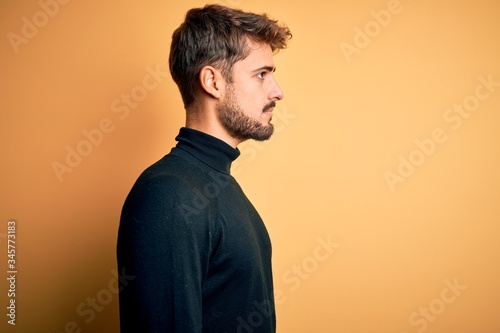 Young handsome man with beard wearing turtleneck sweater standing over yellow background looking to side, relax profile pose with natural face with confident smile.