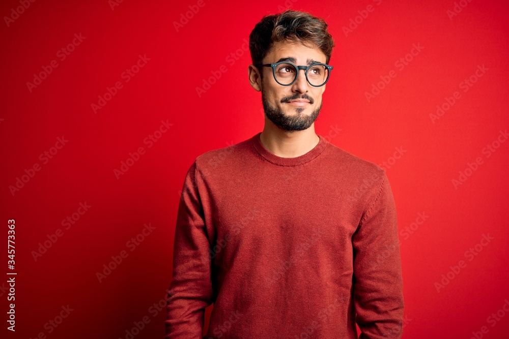 Young handsome man with beard wearing glasses and sweater standing over red background smiling looking to the side and staring away thinking. Young handsome man with beard wearing glasses and sweater standing over red background smiling looking to the side and staring away thinking.