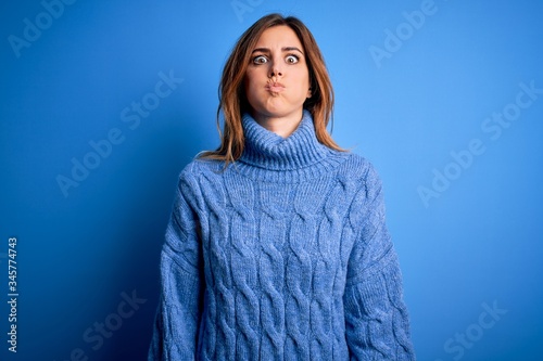 Young beautiful brunette woman wearing casual turtleneck sweater over blue background puffing cheeks with funny face. Mouth inflated with air, crazy expression. photo