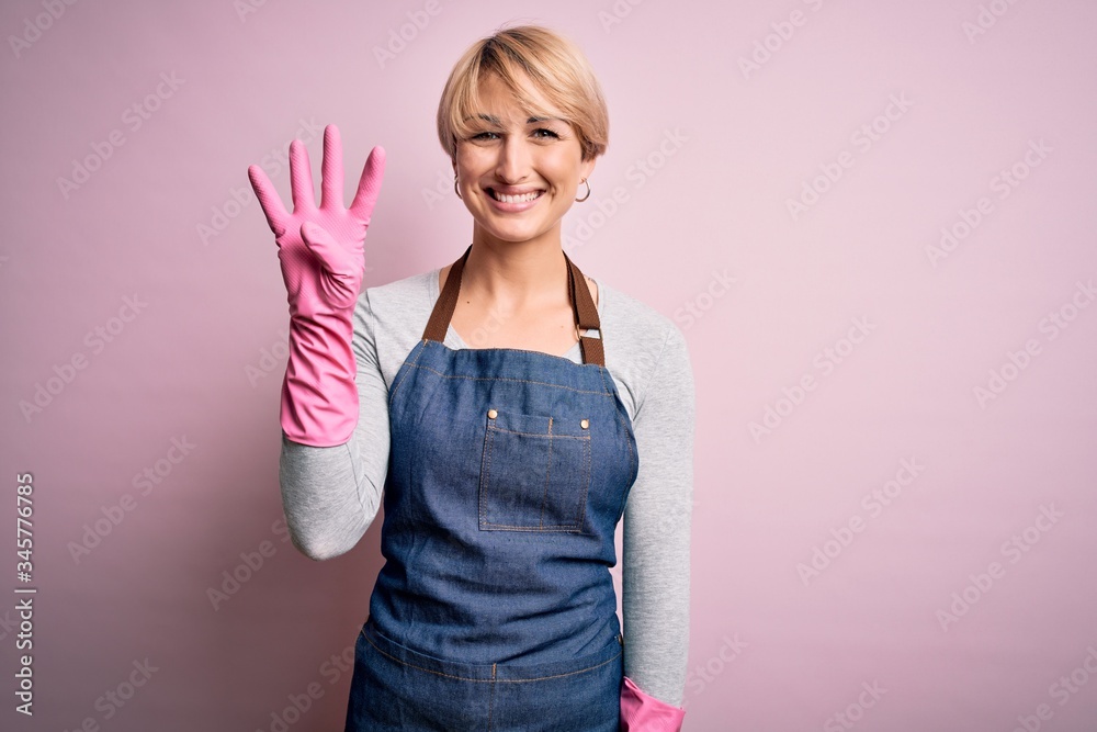 Young blonde cleaner woman with short hair wearing apron and gloves over pink background showing and pointing up with fingers number four while smiling confident and happy. Young blonde cleaner woman with short hair wearing apron and gloves over pink background showing and pointing up with fingers number four while smiling confident and happy.