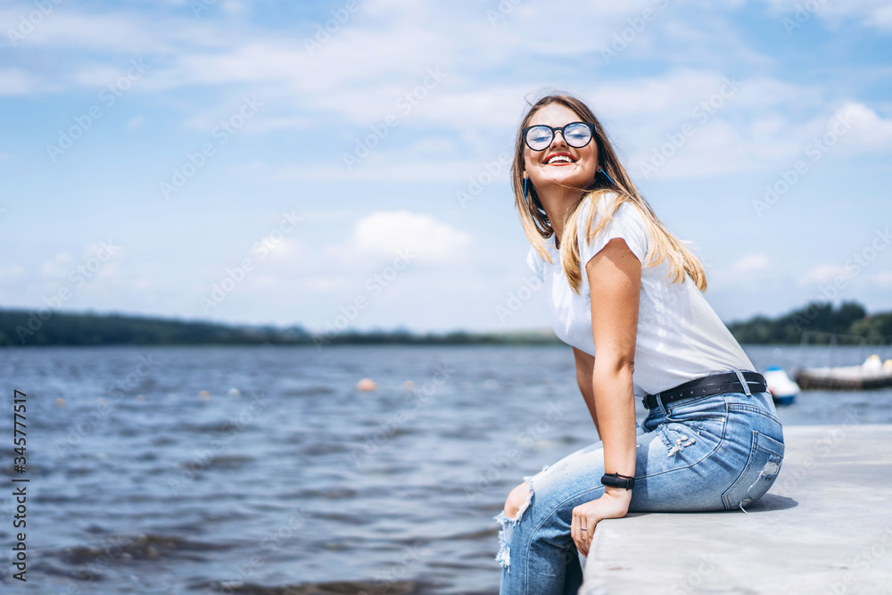 Young woman with long hair in stylish glasses posing on the concrete shore near the lake. Girl dressed in jeans and t-shirt smiling and looking at the camera Young woman with long hair in stylish glasses posing on the concrete shore near the lake. Girl dressed in jeans and t-shirt smiling and looking at the camera