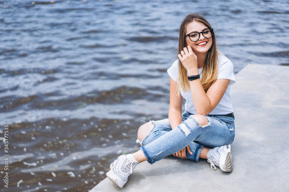 Young woman with long hair in stylish glasses posing on the concrete shore near the lake. Girl dressed in jeans and t-shirt smiling and looking at the camera Young woman with long hair in stylish glasses posing on the concrete shore near the lake. Girl dressed in jeans and t-shirt smiling and looking at the camera