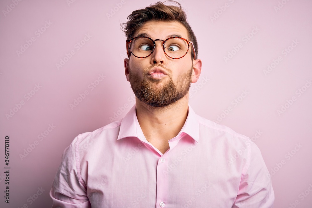 Young handsome blond man with beard and blue eyes wearing pink shirt and glasses making fish face with lips, crazy and comical gesture. Funny expression. Young handsome blond man with beard and blue eyes wearing pink shirt and glasses making fish face with lips, crazy and comical gesture. Funny expression.