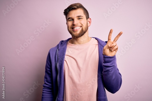 Young blond man with beard and blue eyes wearing purple sweatshirt over pink background showing and pointing up with fingers number two while smiling confident and happy.