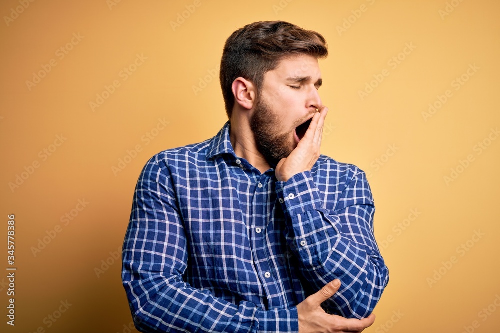 Young blond businessman with beard and blue eyes wearing shirt over yellow background bored yawning tired covering mouth with hand. Restless and sleepiness.
