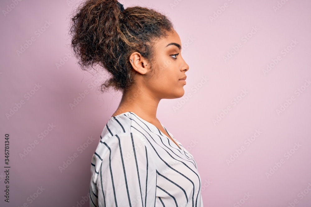 Young beautiful african american girl wearing striped t-shirt standing over pink background looking to side, relax profile pose with natural face with confident smile. Young beautiful african american girl wearing striped t-shirt standing over pink background looking to side, relax profile pose with natural face with confident smile.