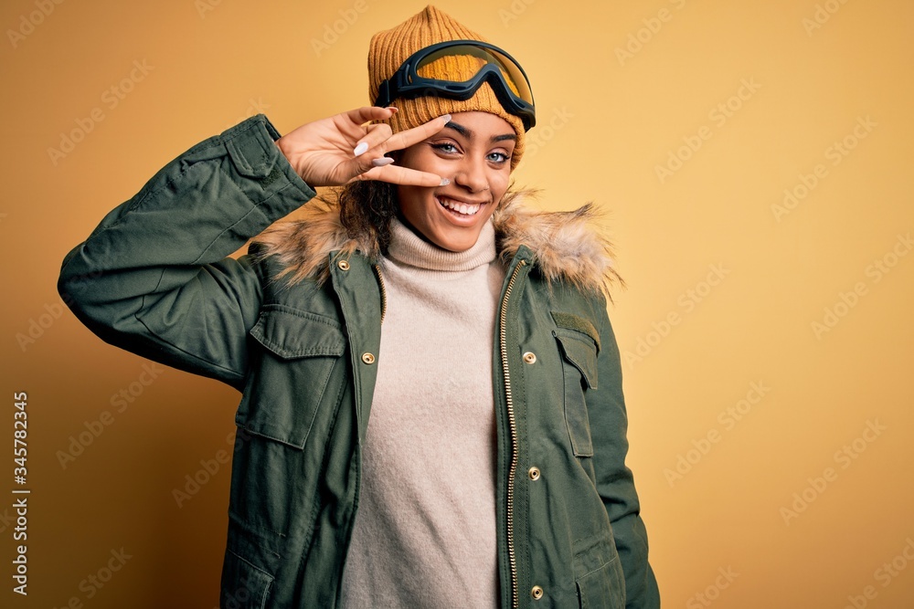 Young african american afro skier girl wearing snow sportswear and ski goggles Doing peace symbol with fingers over face, smiling cheerful showing victory Young african american afro skier girl wearing snow sportswear and ski goggles Doing peace symbol with fingers over face, smiling cheerful showing victory