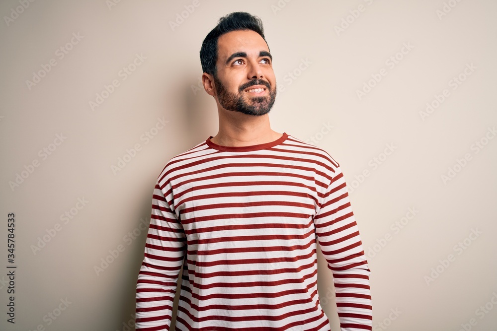 Young handsome man with beard wearing casual striped t-shirt standing over white background looking away to side with smile on face, natural expression. Laughing confident. Young handsome man with beard wearing casual striped t-shirt standing over white background looking away to side with smile on face, natural expression. Laughing confident.