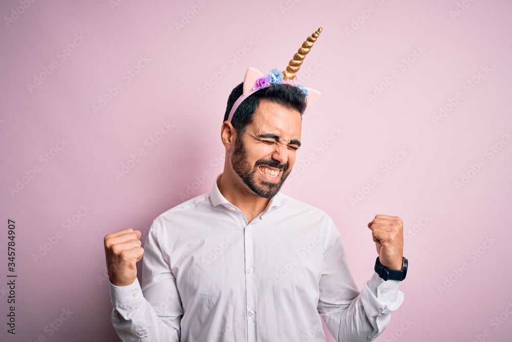 Young handsome man with beard wearing funny unicorn diadem over pink background very happy and excited doing winner gesture with arms raised, smiling and screaming for success. Celebration concept.