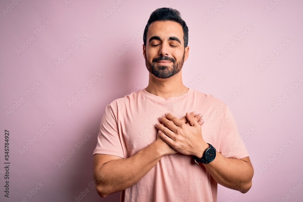 Young handsome man with beard wearing casual t-shirt standing over pink background smiling with hands on chest with closed eyes and grateful gesture on face. Health concept.