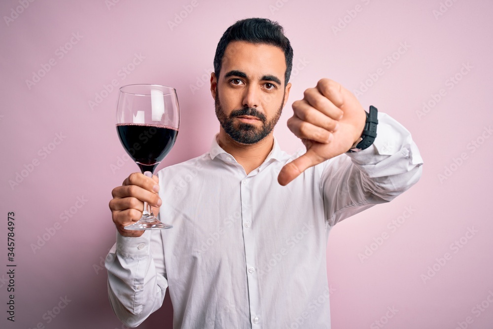 Young handsome man with beard drinking glass of red wine over isolated pink background with angry face, negative sign showing dislike with thumbs down, rejection concept Young handsome man with beard drinking glass of red wine over isolated pink background with angry face, negative sign showing dislike with thumbs down, rejection concept