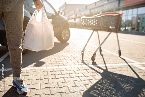 Close-up of a stroller with food near a large supermarket in a suburban shopping center. A man stands near a car in a parking lot after a successful shopping © Vlad