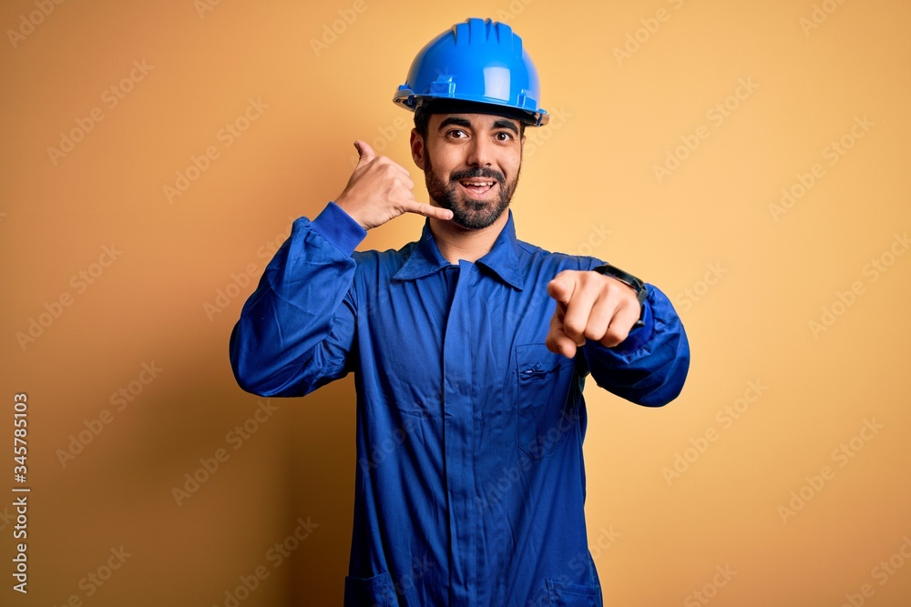 Mechanic man with beard wearing blue uniform and safety helmet over yellow background smiling doing talking on the telephone gesture and pointing to you. Call me. Mechanic man with beard wearing blue uniform and safety helmet over yellow background smiling doing talking on the telephone gesture and pointing to you. Call me.