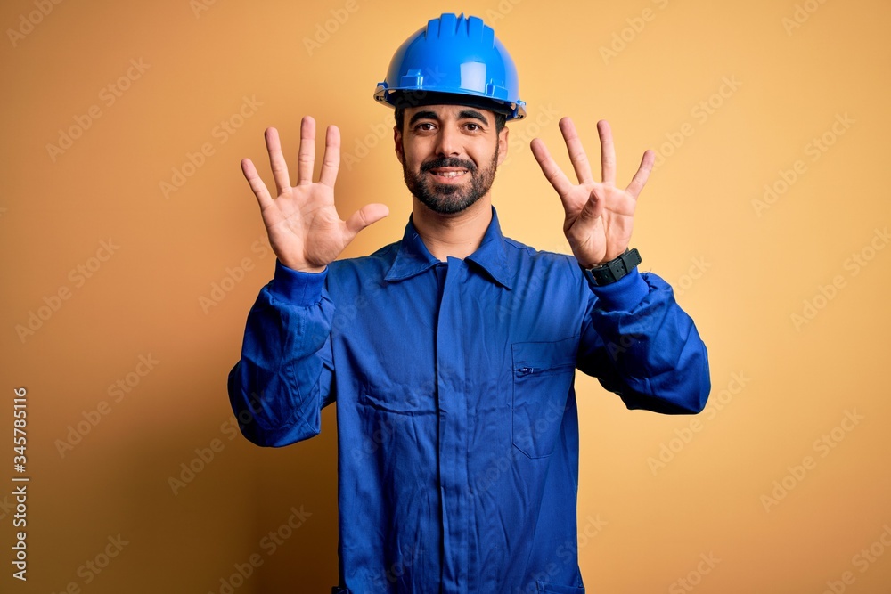 Mechanic man with beard wearing blue uniform and safety helmet over yellow background showing and pointing up with fingers number nine while smiling confident and happy.