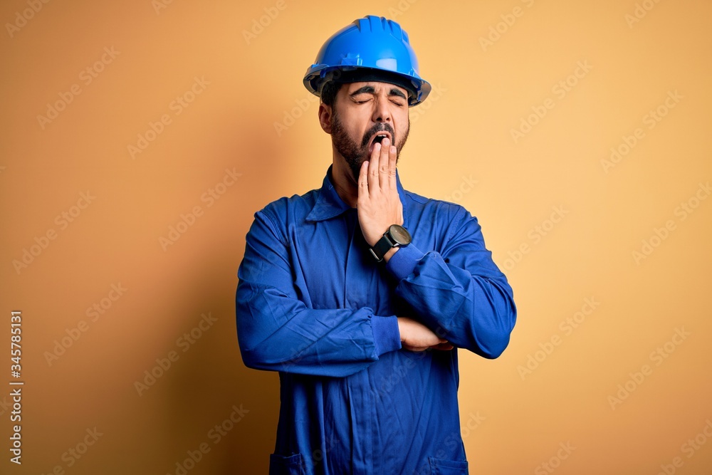 Mechanic man with beard wearing blue uniform and safety helmet over yellow background bored yawning tired covering mouth with hand. Restless and sleepiness. Mechanic man with beard wearing blue uniform and safety helmet over yellow background bored yawning tired covering mouth with hand. Restless and sleepiness.