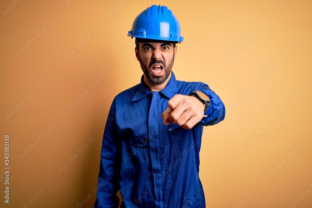 Mechanic man with beard wearing blue uniform and safety helmet over yellow background pointing displeased and frustrated to the camera, angry and furious with you