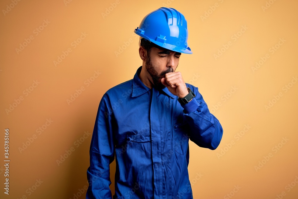 Mechanic man with beard wearing blue uniform and safety helmet over yellow background feeling unwell and coughing as symptom for cold or bronchitis. Health care concept.
