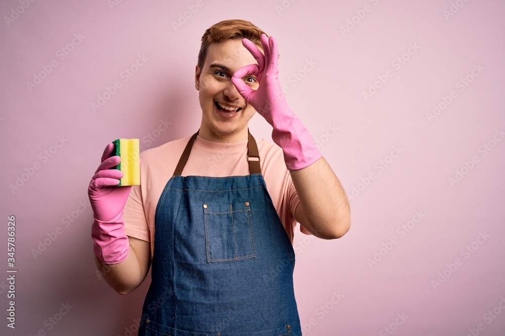 Handsome redhead man doing housework wearing apron and gloves using cleaner scourer smiling happy doing ok sign with hand on eye looking through fingers Handsome redhead man doing housework wearing apron and gloves using cleaner scourer smiling happy doing ok sign with hand on eye looking through fingers