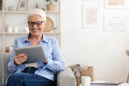 Smiling elderly lady wearing glasses using digital tablet at home