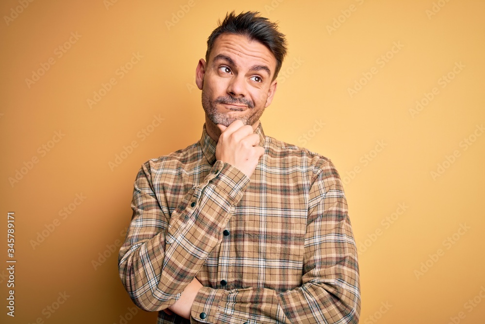 Young handsome man wearing casual shirt standing over isolated yellow background with hand on chin thinking about question, pensive expression. Smiling with thoughtful face. Doubt concept. Young handsome man wearing casual shirt standing over isolated yellow background with hand on chin thinking about question, pensive expression. Smiling with thoughtful face. Doubt concept.