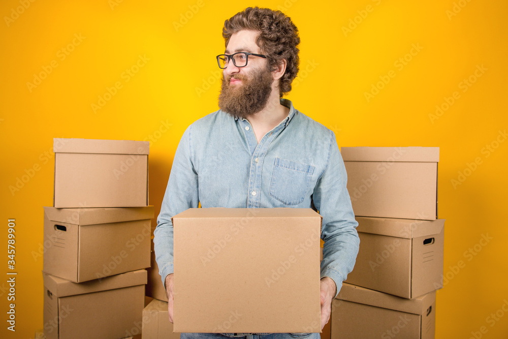 Bearded man with glasses a delivery man holding a cardboard box in his hands standing among many boxes with parcels on a yellow background Bearded man with glasses a delivery man holding a cardboard box in his hands standing among many boxes with parcels on a yellow background