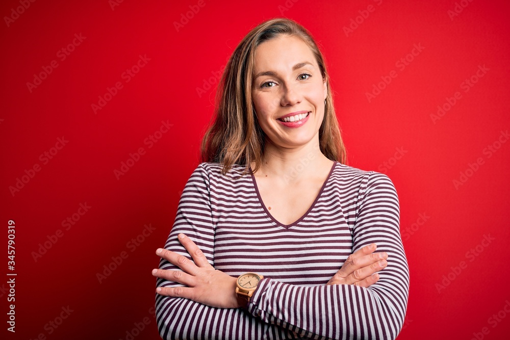Young beautiful blonde woman wearing casual striped t-shirt over isolated red background happy face smiling with crossed arms looking at the camera. Positive person. Young beautiful blonde woman wearing casual striped t-shirt over isolated red background happy face smiling with crossed arms looking at the camera. Positive person.