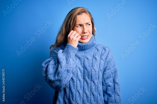 Young beautiful blonde woman wearing casual turtleneck sweater over blue background looking stressed and nervous with hands on mouth biting nails. Anxiety problem.