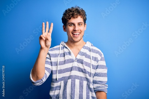Young blond handsome man with curly hair wearing casual striped sweatshirt showing and pointing up with fingers number three while smiling confident and happy.