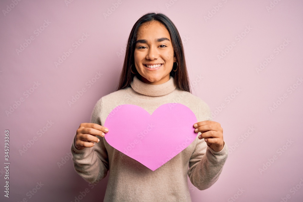 Young beautiful asian woman holding pink heart standing over isolated background with a happy face standing and smiling with a confident smile showing teeth Young beautiful asian woman holding pink heart standing over isolated background with a happy face standing and smiling with a confident smile showing teeth