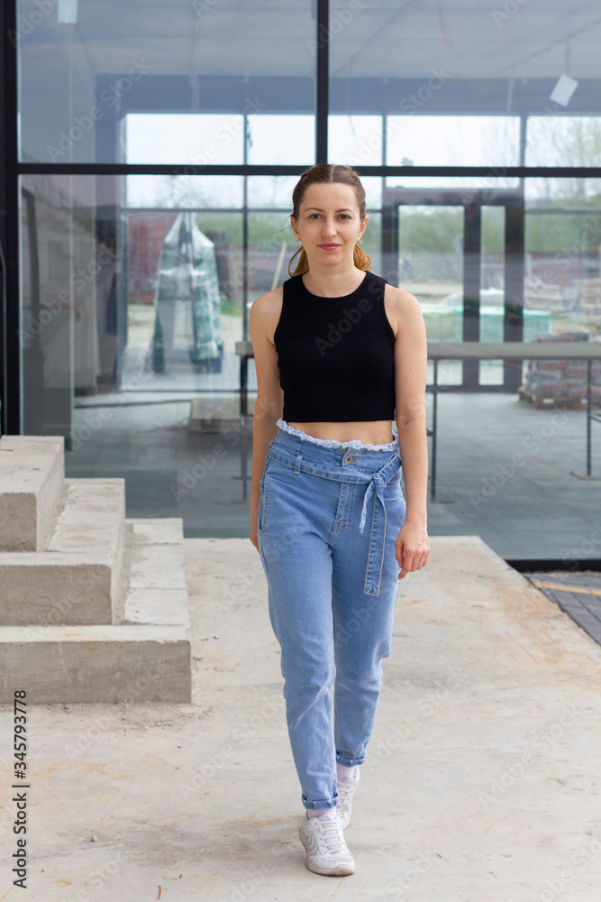 Gorgeous fashion sexy female dressed in black cropp top and blue jeans standing outdoor, smiling and enjoying sunny day. Sports arena in the background. Girl posing at sunny day.