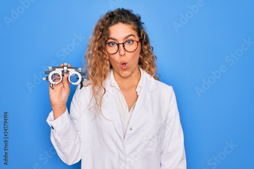 Beautiful oculist woman with blue eyes holding optometry glasses over isolated background scared in shock with a surprise face, afraid and excited with fear expression