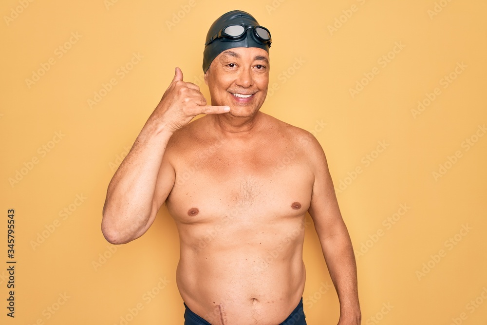 Middle age senior grey-haired swimmer man wearing swimsuit, cap and goggles smiling doing phone gesture with hand and fingers like talking on the telephone. Communicating concepts.