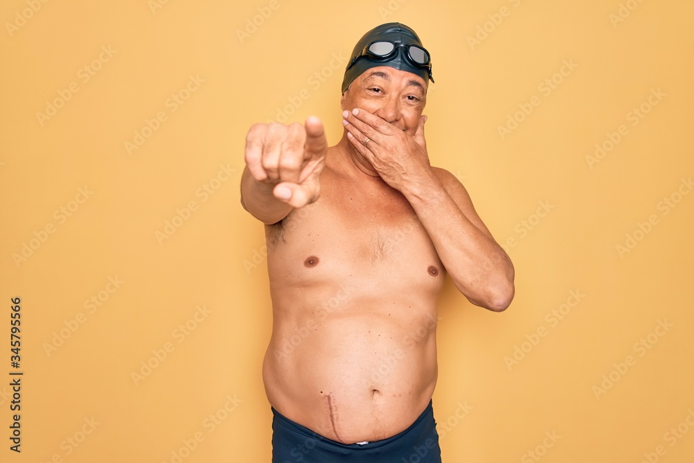Middle age senior grey-haired swimmer man wearing swimsuit, cap and goggles laughing at you, pointing finger to the camera with hand over mouth, shame expression