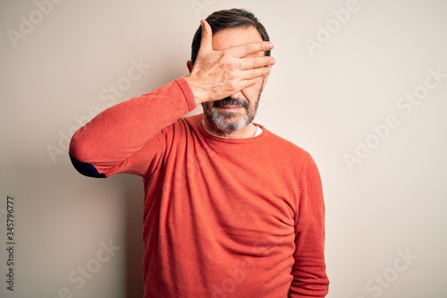 Middle age hoary man wearing casual orange sweater standing over isolated white background covering eyes with hand, looking serious and sad. Sightless, hiding and rejection concept