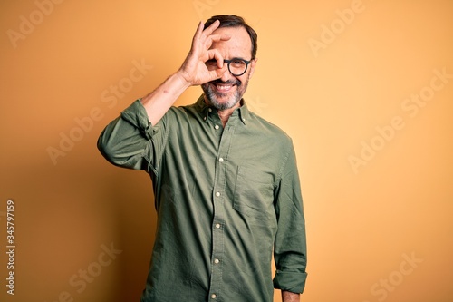 Middle age hoary man wearing casual green shirt and glasses over isolated yellow background doing ok gesture with hand smiling, eye looking through fingers with happy face. © Krakenimages.com