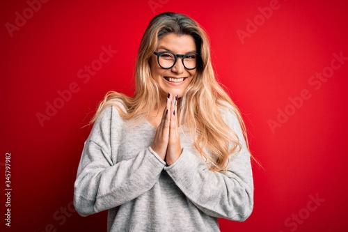 Young beautiful blonde woman wearing sweater and glasses over isolated red background praying with hands together asking for forgiveness smiling confident.