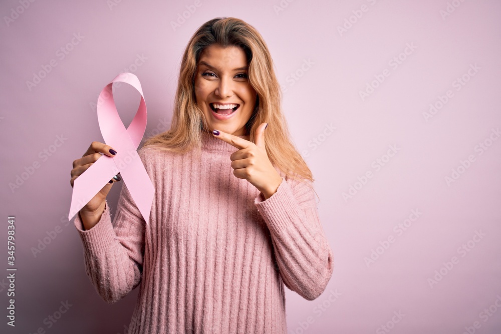 Young beautiful blonde woman asking for support cancer holding pink ribbon symbol very happy pointing with hand and finger Young beautiful blonde woman asking for support cancer holding pink ribbon symbol very happy pointing with hand and finger