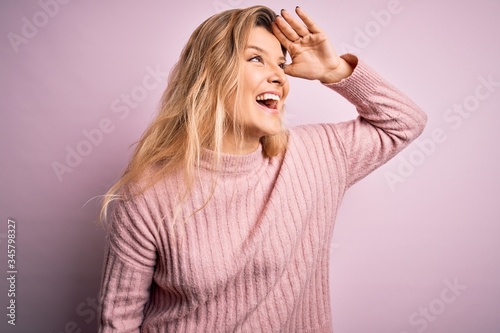 Young beautiful blonde woman wearing casual pink sweater over isolated background very happy and smiling looking far away with hand over head. Searching concept.