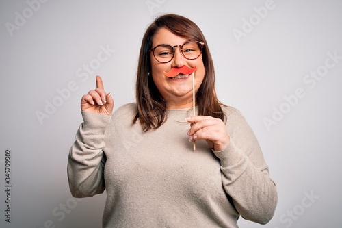 Beautiful plus size woman holding fake party mustache over isolated background surprised with an idea or question pointing finger with happy face  number one