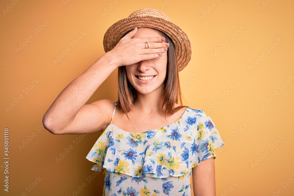 Young beautiful woman wearing casual t-shirt and summer hat over isolated yellow background smiling and laughing with hand on face covering eyes for surprise. Blind concept. Young beautiful woman wearing casual t-shirt and summer hat over isolated yellow background smiling and laughing with hand on face covering eyes for surprise. Blind concept.
