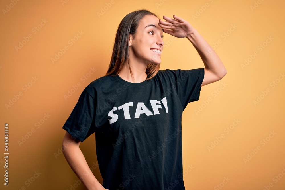 Young beautiful worker woman wearing staff uniform t-shirt over isolated yellow background very happy and smiling looking far away with hand over head. Searching concept. Young beautiful worker woman wearing staff uniform t-shirt over isolated yellow background very happy and smiling looking far away with hand over head. Searching concept.