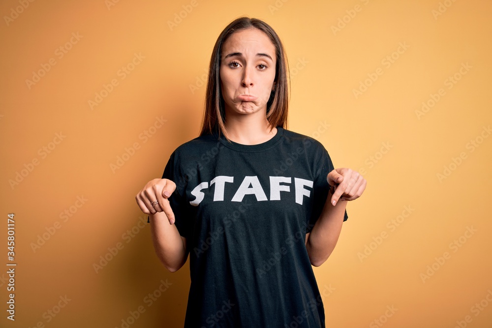 Young beautiful worker woman wearing staff uniform t-shirt over isolated yellow background Pointing down looking sad and upset, indicating direction with fingers, unhappy and depressed. Young beautiful worker woman wearing staff uniform t-shirt over isolated yellow background Pointing down looking sad and upset, indicating direction with fingers, unhappy and depressed.