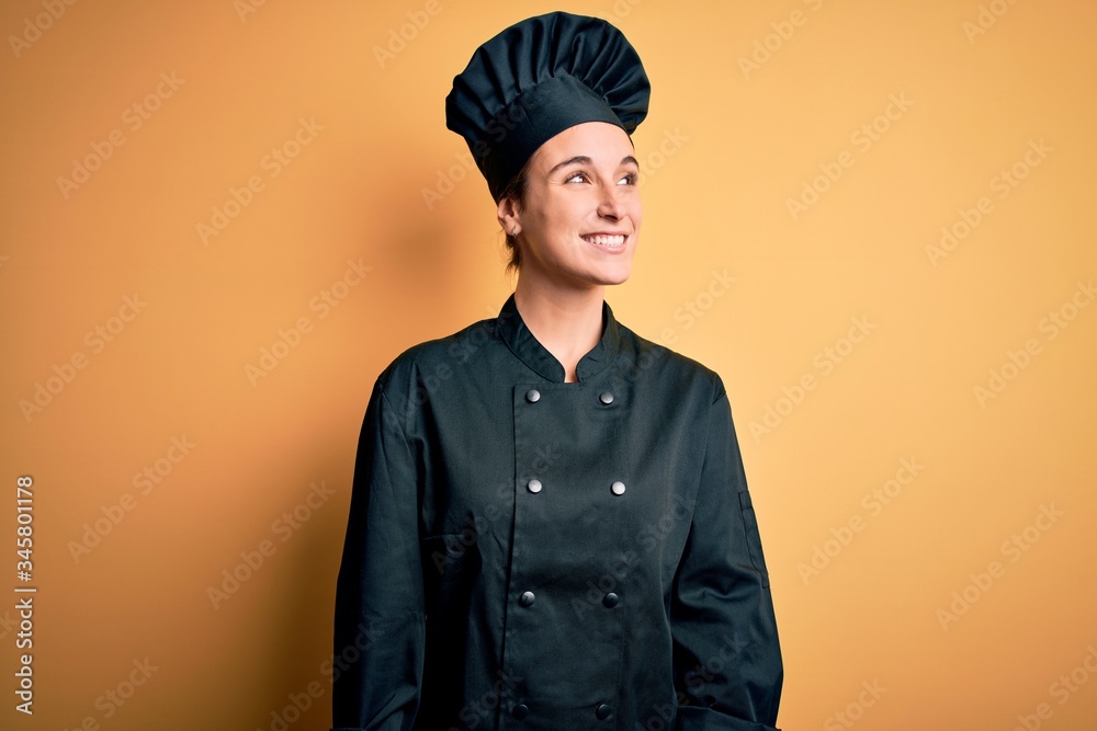 Young beautiful chef woman wearing cooker uniform and hat standing over yellow background looking away to side with smile on face, natural expression. Laughing confident. Young beautiful chef woman wearing cooker uniform and hat standing over yellow background looking away to side with smile on face, natural expression. Laughing confident.