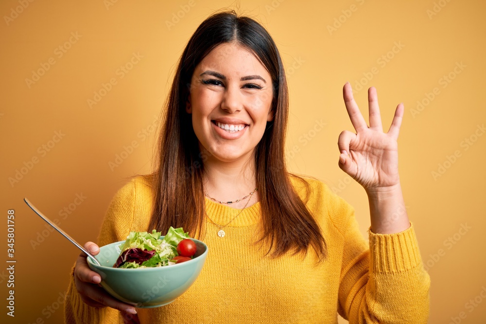 Young beautiful woman eating healthy fresh salad over yellow background doing ok sign with fingers, excellent symbol Young beautiful woman eating healthy fresh salad over yellow background doing ok sign with fingers, excellent symbol