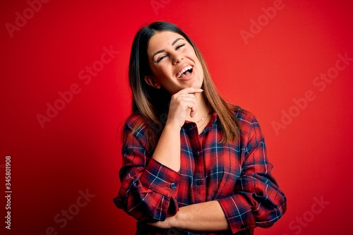 Young beautiful woman wearing casual shirt over red background looking confident at the camera smiling with crossed arms and hand raised on chin. Thinking positive. © Krakenimages.com
