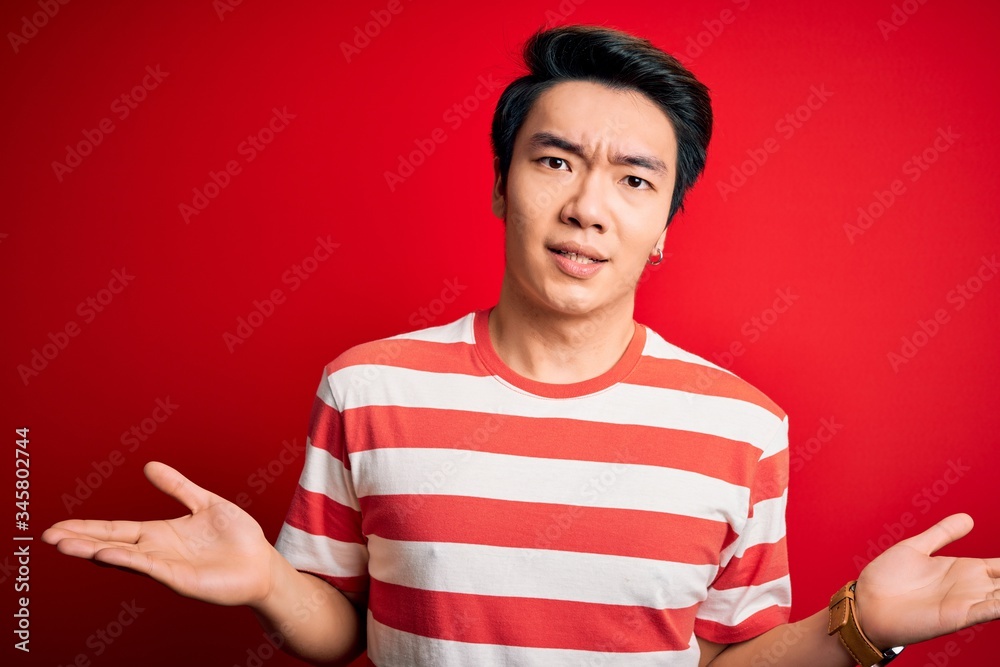Young handsome chinese man wearing casual striped t-shirt standing over red background clueless and confused with open arms, no idea concept.